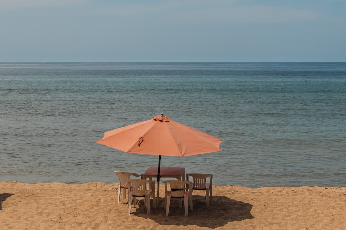 A tranquil beach setting featuring an orange umbrella shading a small table with four white chairs on the golden sand, overlooking the vast blue ocean, emphasizing the property's direct beach access.