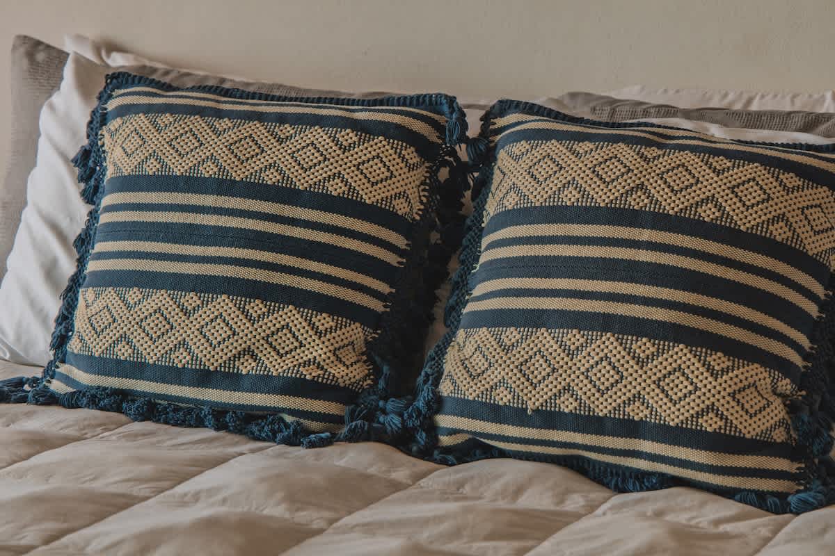 Two rectangular decorative pillows with a beige and blue striped pattern and diamond-textured accents, featuring blue tassel trim, rest on a light-colored quilted bedspread.