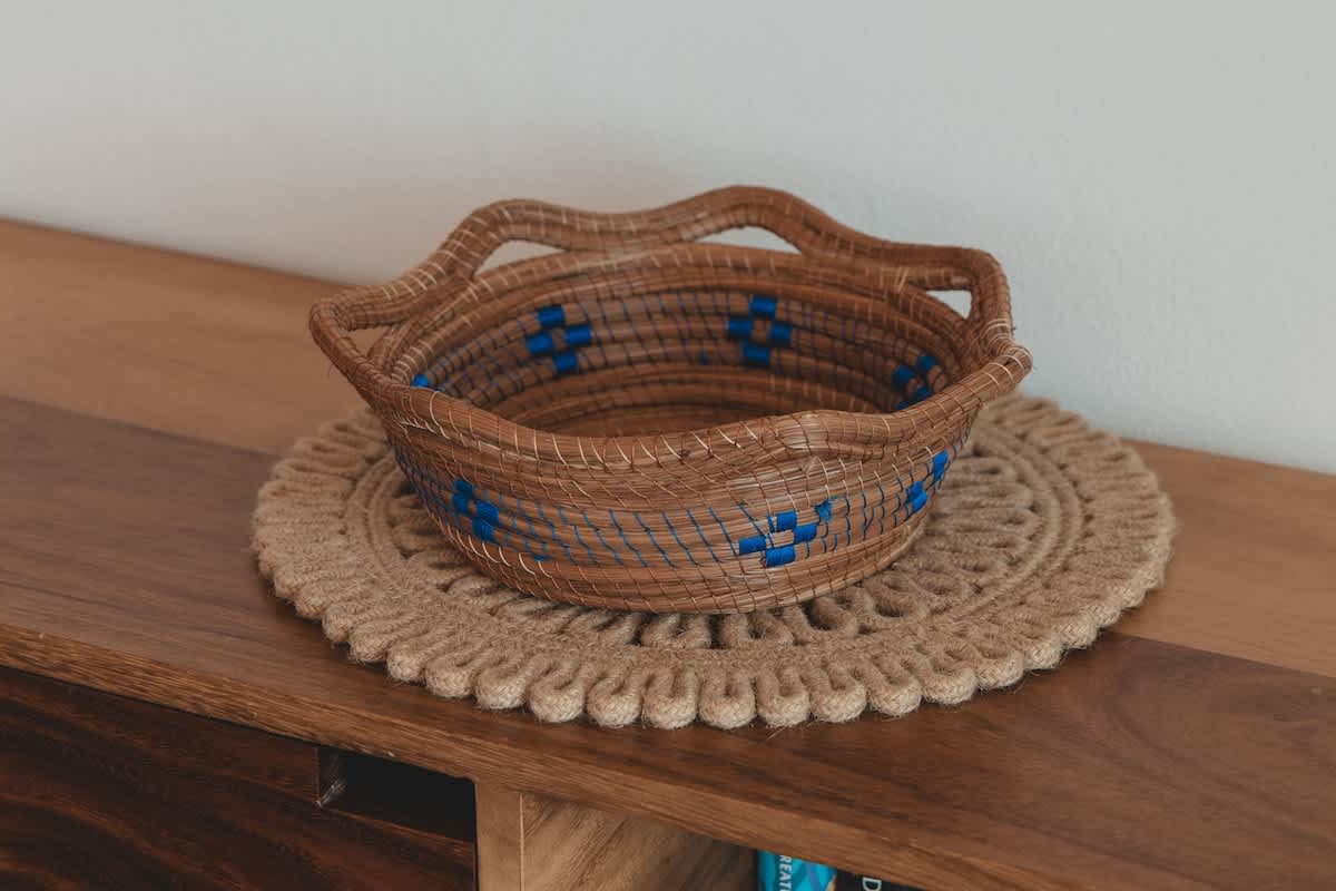 A rustic woven basket with blue accents sits on a round, textured beige placemat, both resting on a polished wooden surface, possibly a console table or dresser.