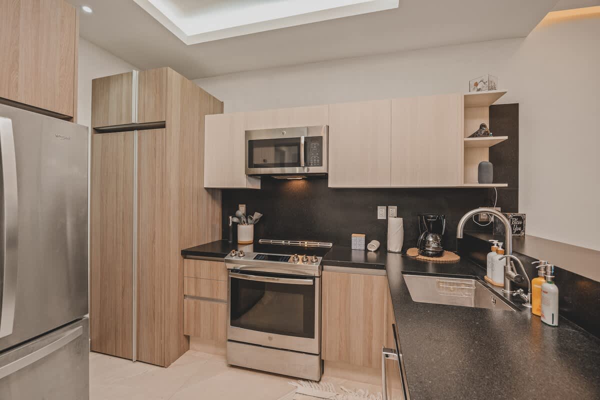 Wide shot of the modern kitchen, featuring stainless steel appliances, light wood cabinetry, dark countertops, and a breakfast bar. The room is illuminated by recessed lighting and a central skylight.