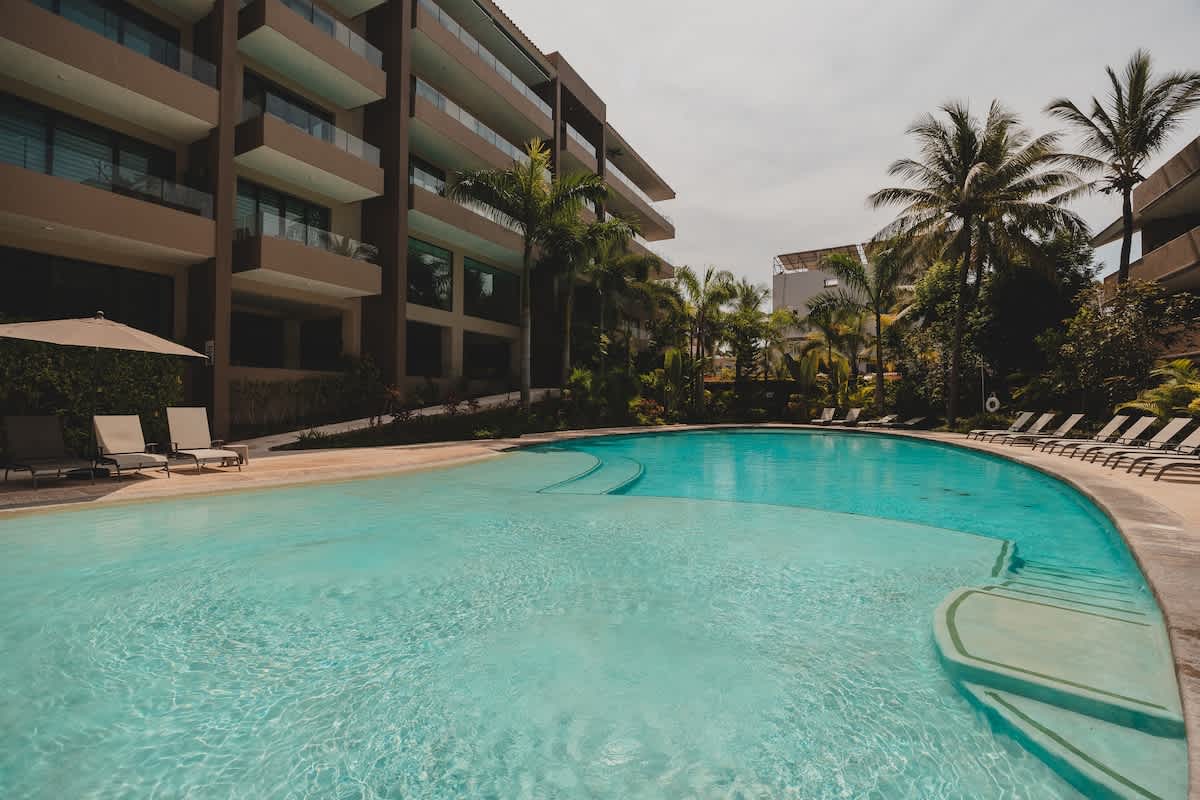 A wide view of a resort-style swimming pool with clear blue water, featuring a shallow entry area and surrounded by numerous lounge chairs. Modern multi-story buildings and palm trees frame the sunny outdoor space.