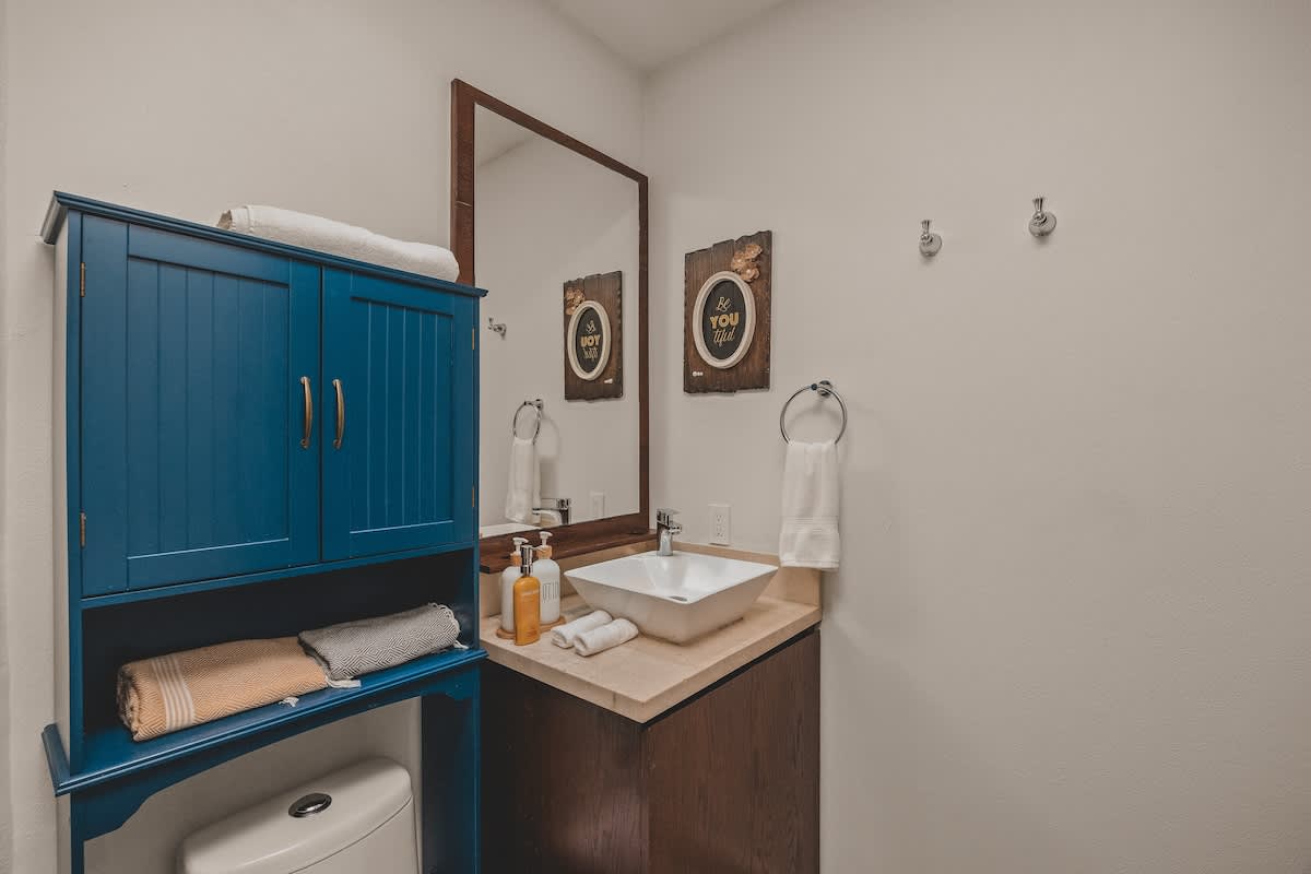 A modern bathroom featuring a dark wood vanity with a white vessel sink, a large mirror, and a blue wall-mounted cabinet above the toilet. White towels and decorative wall art complete the clean aesthetic.