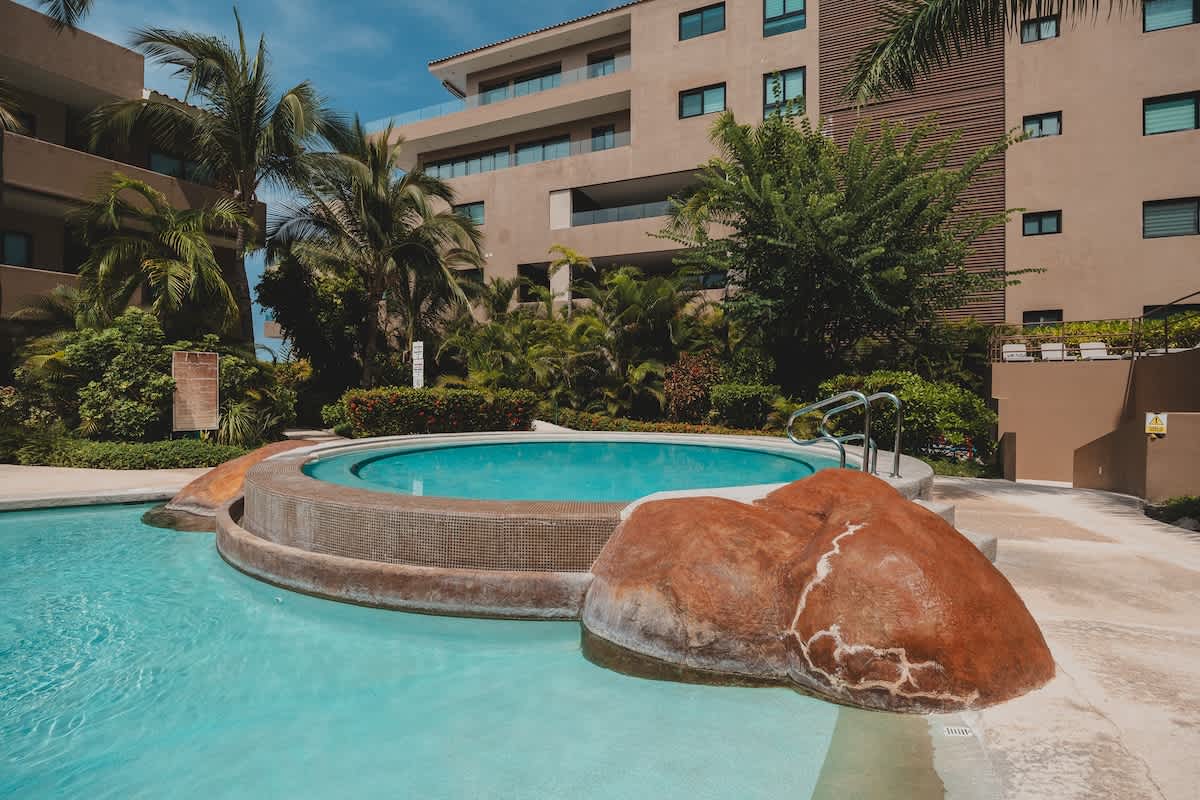 A circular hot tub with tiled sides and decorative rock features, connected to a larger swimming pool, set amidst concrete decking and vibrant tropical plants under a clear sky.