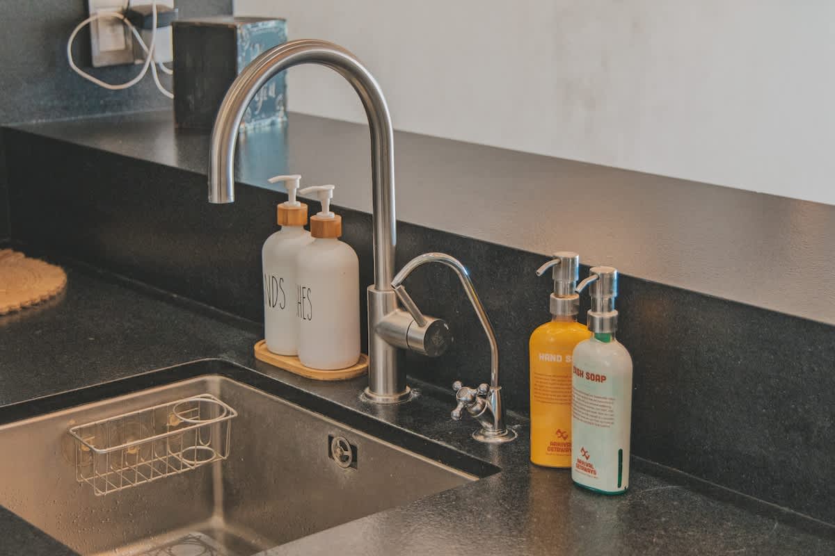 A close-up of a modern kitchen sink area featuring a gooseneck faucet, a smaller water filter faucet, and multiple soap dispensers on a dark, sleek countertop. A metal sponge holder is visible in the stainless steel sink.