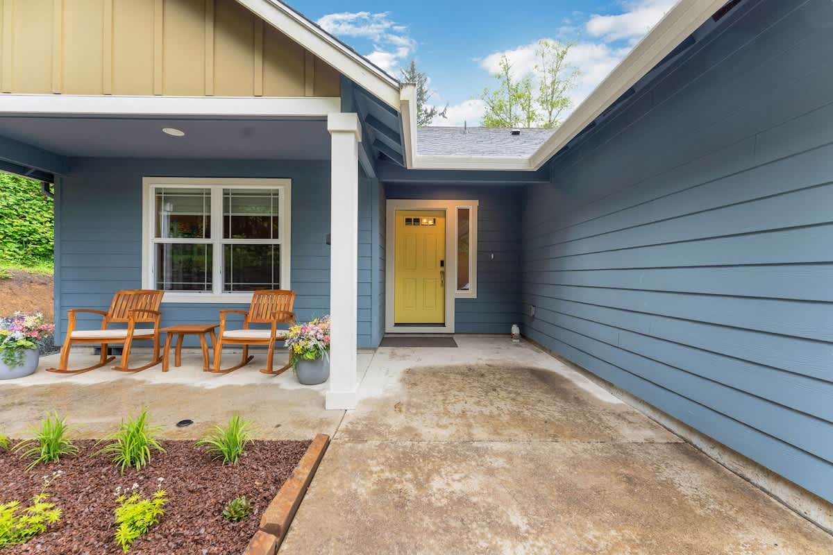 A close-up of the covered front porch with a bright yellow front door, flanked by a window and white support columns. Two wooden rocking chairs and a small table are arranged on the concrete porch, with landscaped planting beds nearby.