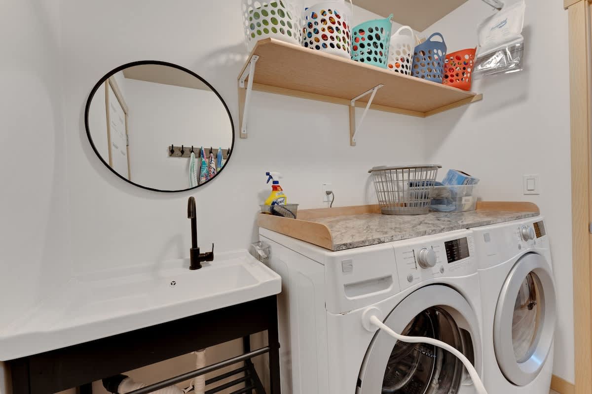A functional laundry room features a white front-loading washer and dryer with a light countertop above, and a white utility sink with a black faucet. A round black mirror hangs on the wall, and a wood shelf above the machines holds colorful laundry baskets.