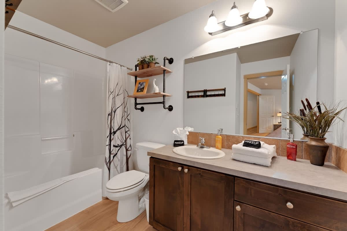 A bright bathroom features a dark wood single vanity with a light countertop and a white sink, topped with a large mirror and a three-light fixture. A white bathtub with a shower and a toilet are on the left, adorned with a tree-patterned shower curtain and industrial-style shelving above the toilet.