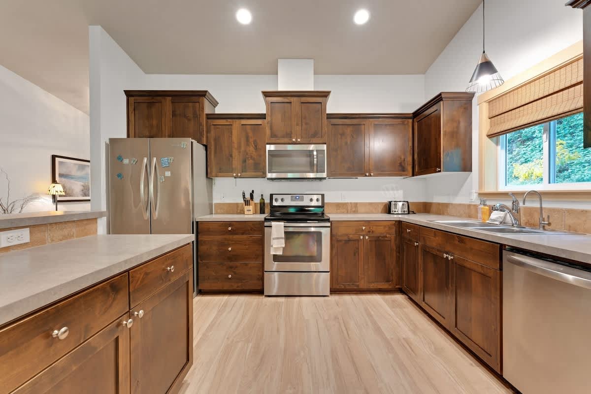 A spacious kitchen with light wood-look flooring, dark wood shaker-style cabinetry, and light beige countertops. Stainless steel appliances, including a refrigerator, oven/range, microwave, and dishwasher, are integrated into the design.