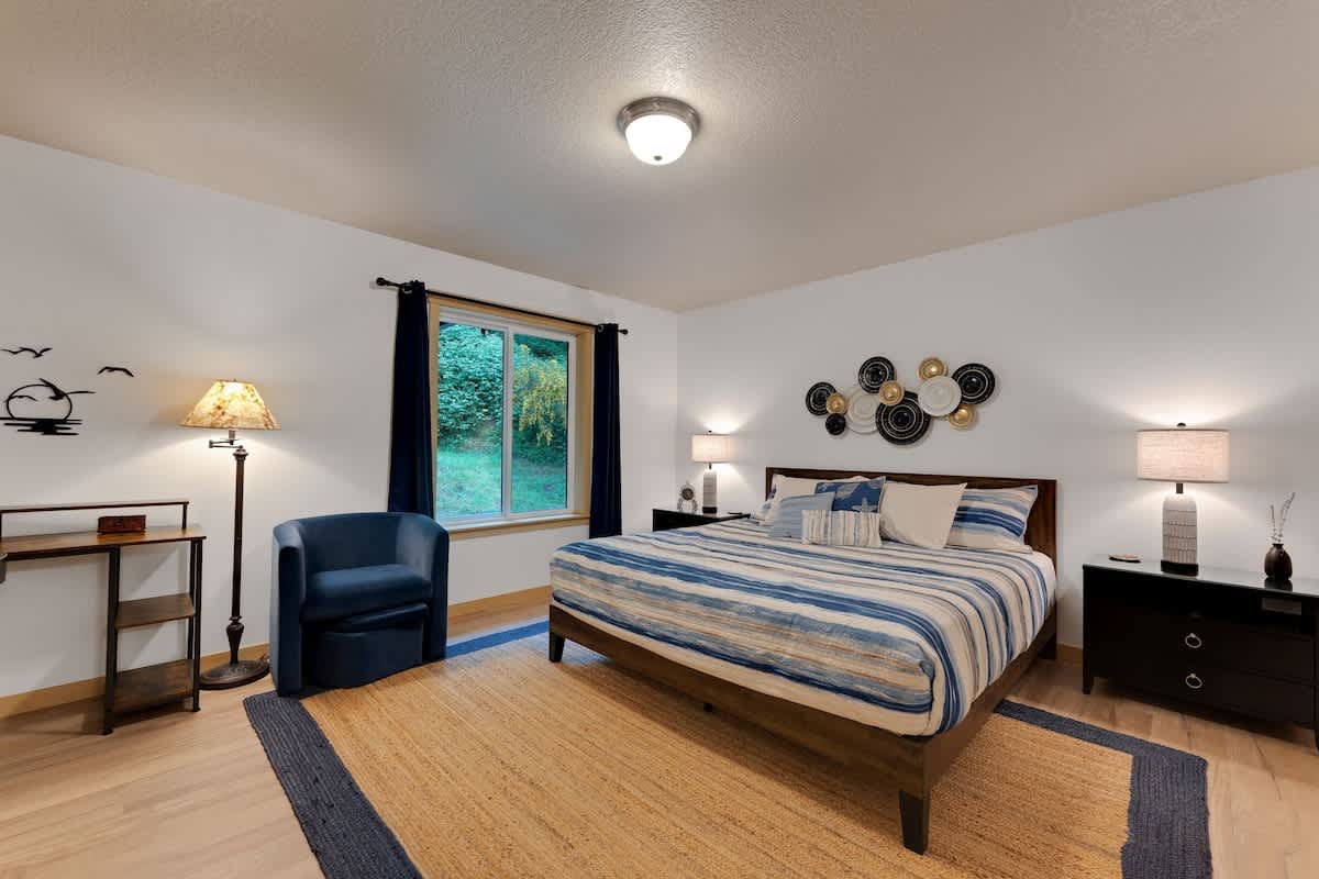 Wide shot of the master bedroom, showcasing a king bed with striped bedding, black nightstands, a blue accent chair, a tall floor lamp, and a window with dark blue curtains, all on a large natural fiber rug.
