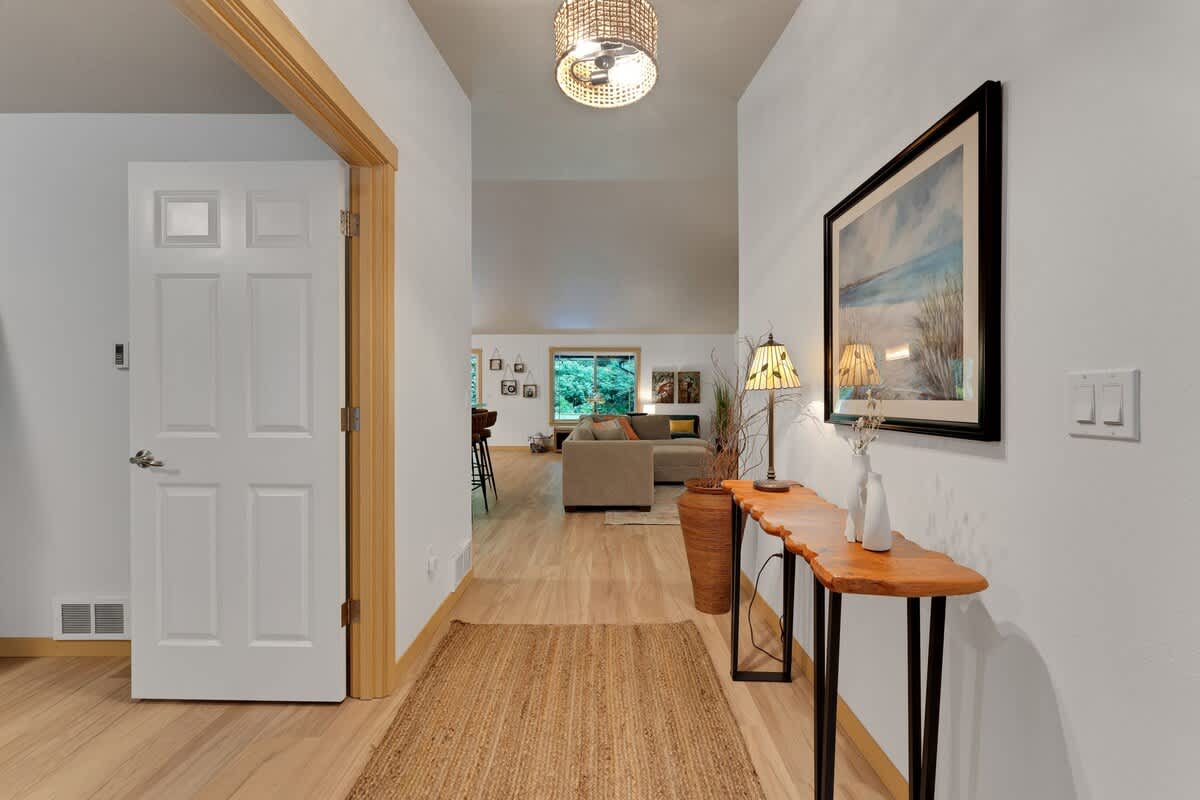 A bright foyer with light hardwood floors and white walls, featuring a decorative console table with a lamp and framed artwork, leading into an open-concept living space.