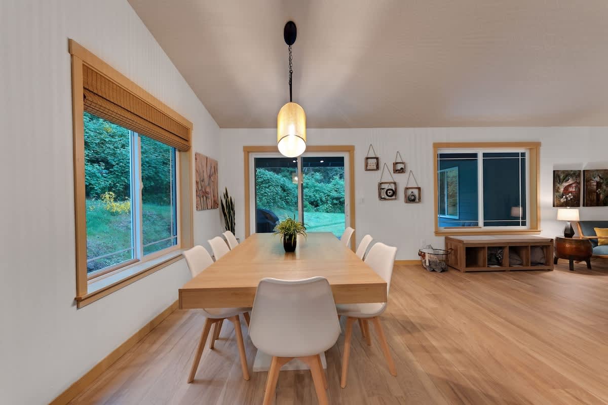 Bright dining area with a long light wood dining table and eight white chairs, illuminated by a modern oval pendant light, with large windows offering views of lush greenery.