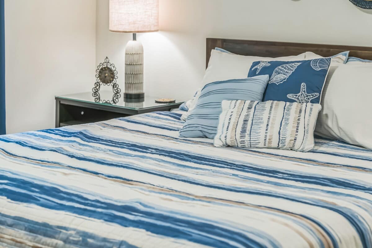 Close-up of a queen-sized bed with a dark wood headboard and blue and white striped bedding, flanked by a glass-top nightstand holding a textured lamp and an antique clock.