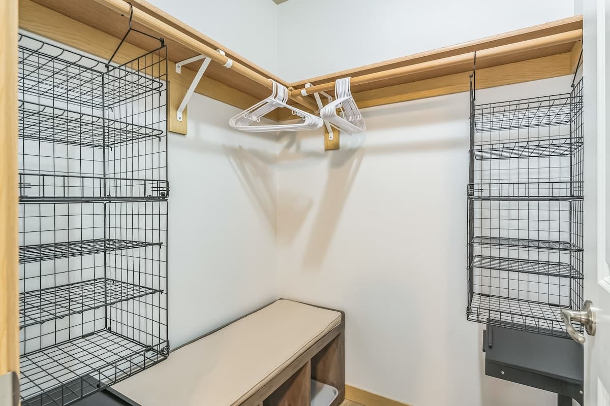 A walk-in closet features light wood shelving and hanging rods, with black wire basket organizers on both sides. A built-in bench with a light cushion provides seating in the center.