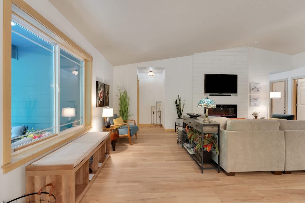 Spacious living area with light hardwood floors, a large window bench, a comfortable armchair, and a view towards a shiplap-accented wall with a fireplace and TV.