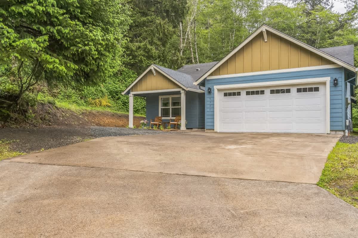A wide concrete driveway extends in front of a blue house with a white two-car garage door and tan gables, set against a backdrop of dense green trees. A small covered porch with outdoor seating is visible on the left.