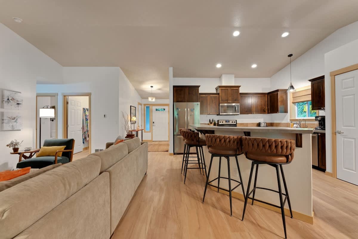 A modern kitchen with dark wood cabinets, stainless steel appliances, and a breakfast bar with three brown leather bar stools, opening into a living area and showing the front door in the distance.