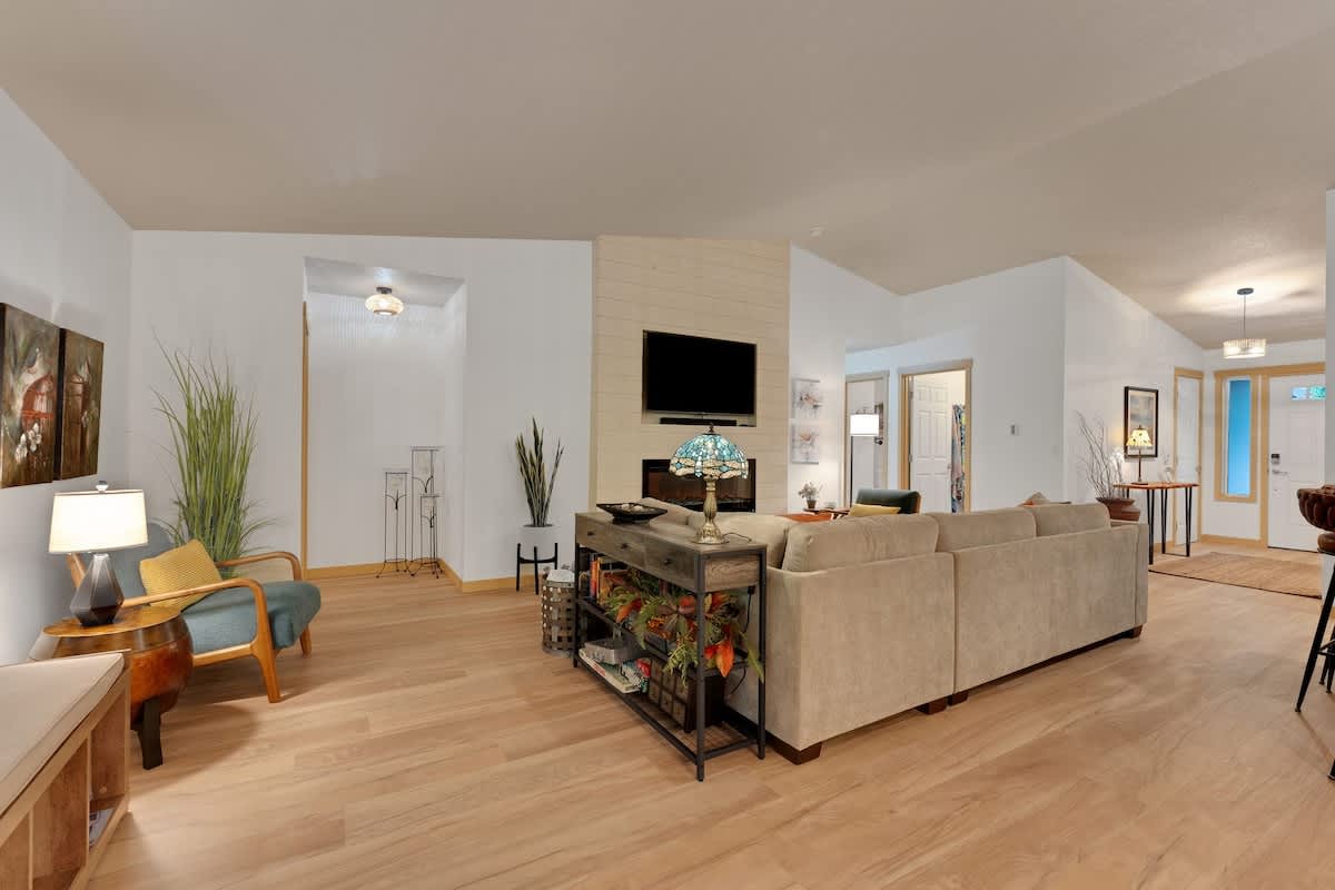 A spacious living room with light hardwood floors and white walls, featuring a large sectional sofa, a fireplace with a mounted TV, and an accent chair, all under a vaulted ceiling.