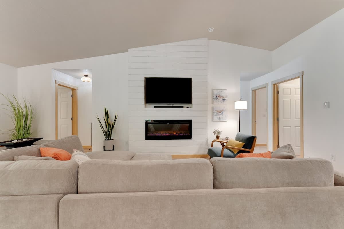Living room with a large sectional sofa facing a shiplap-paneled accent wall, featuring a wall-mounted TV above an electric fireplace, flanked by tall decorative plants.