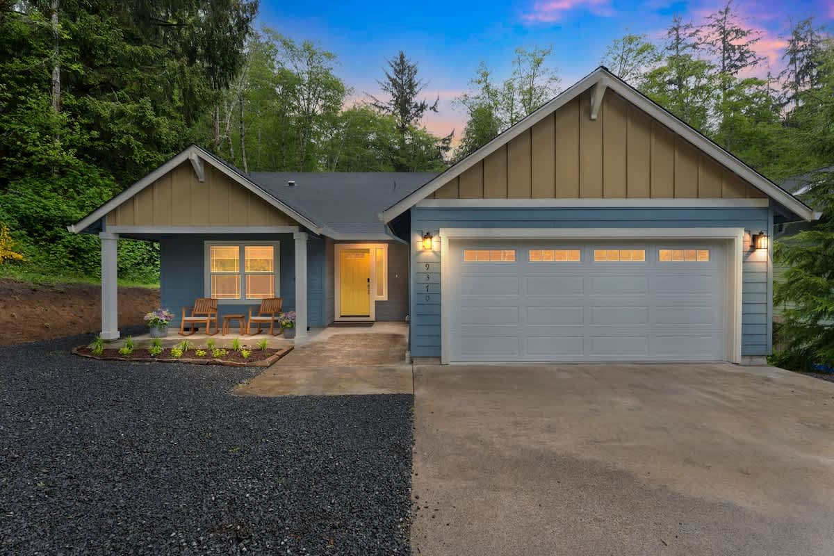The front exterior of a light blue house with a yellow front door and a white garage door, featuring a small covered porch with wooden chairs and a gravel driveway.