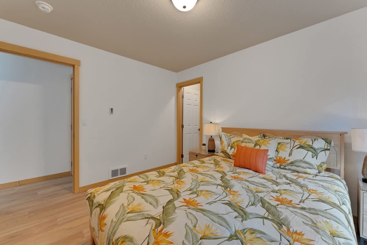 Another view of the bedroom with a queen bed and tropical floral bedding, showing light wood floors, a nightstand, and an open doorway leading to a closet or hallway, with a tall decorative vase.
