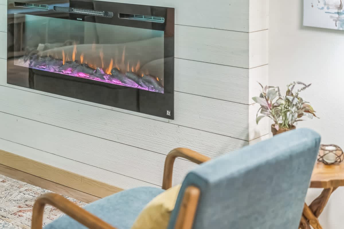 A modern electric fireplace with vibrant, colorful flames is mounted on a white shiplap accent wall, creating a cozy focal point. A light blue armchair with wooden arms and a small rustic wooden side table with a plant are visible in the foreground.