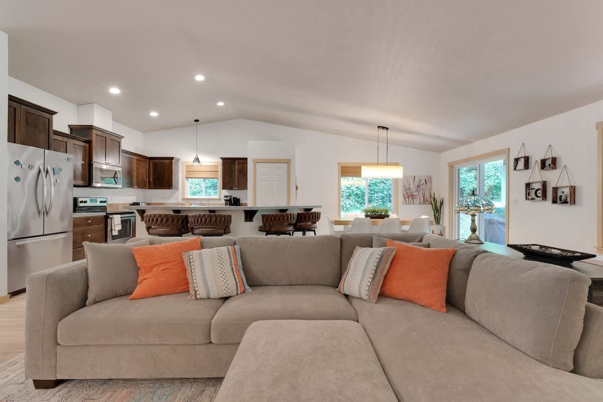 Open-concept living room featuring a large L-shaped sectional sofa with orange and patterned throw pillows, facing an entertainment area, with a view into the kitchen and dining space.