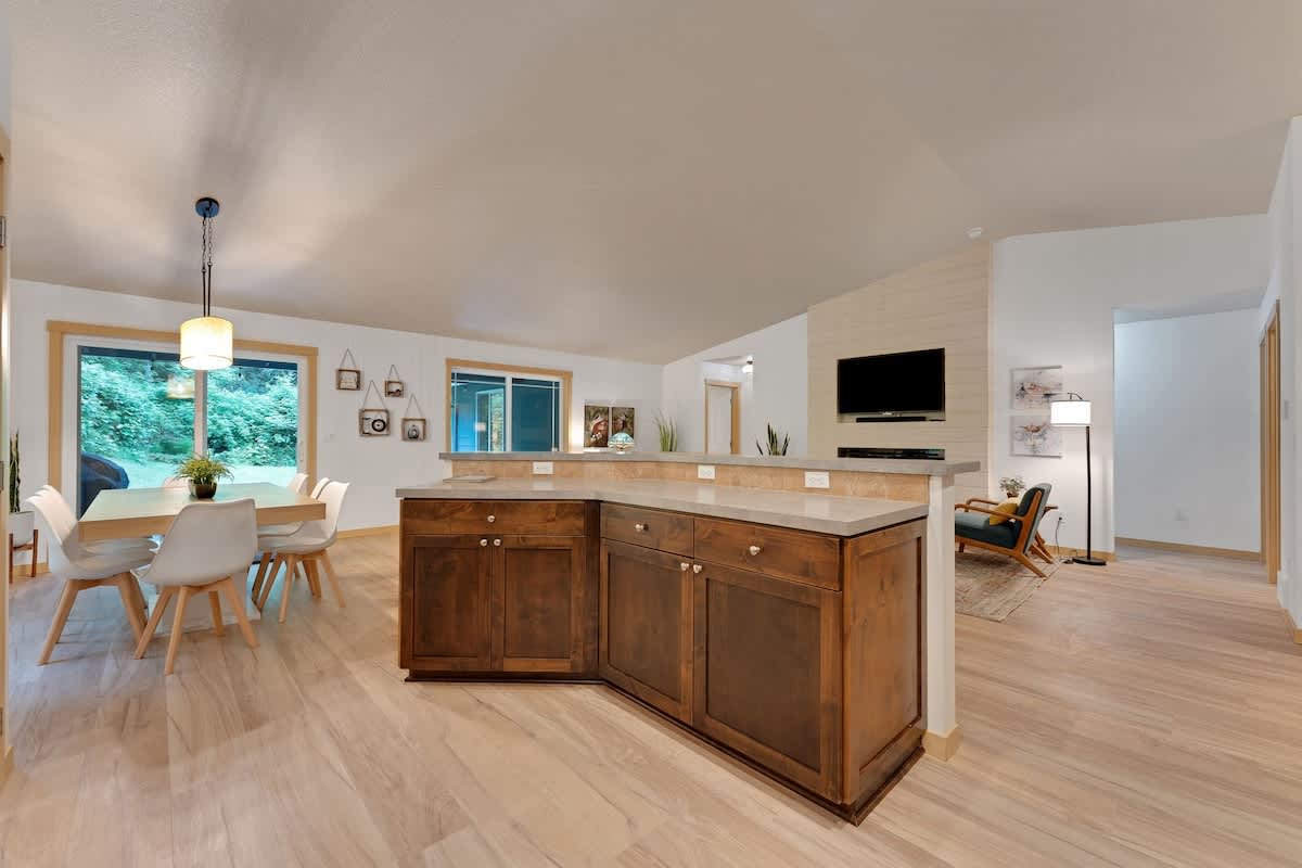 An open-concept dining area with a light wood table and modern white chairs, adjacent to a kitchen island with dark wood cabinetry and light countertops, all on light hardwood floors.