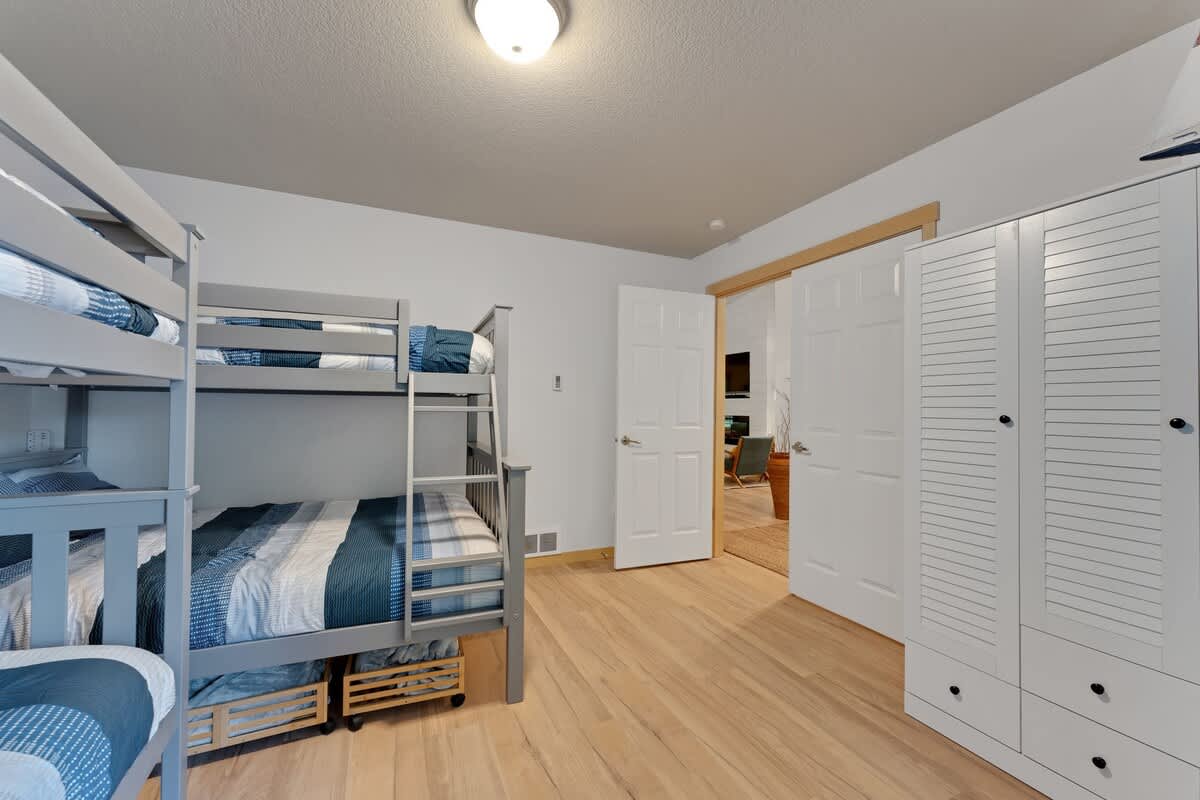 Another view of the bunk room, showing two grey bunk beds with striped bedding, light wood floors, a white wardrobe with louvered doors, and a white door leading to an adjacent room.
