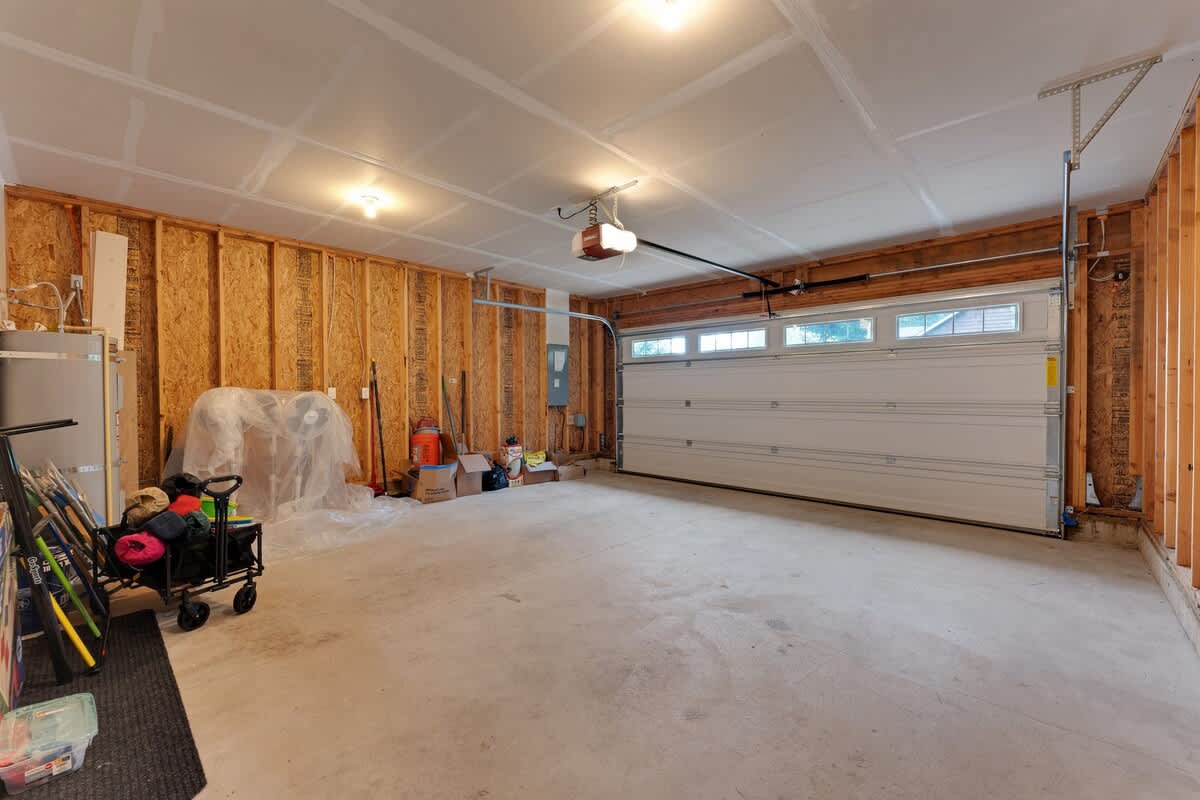 A spacious, unfinished garage features exposed wooden studs on the walls and a concrete floor, with a large white garage door at the far end. Various tools, boxes, and a wagon are stored along the left wall.