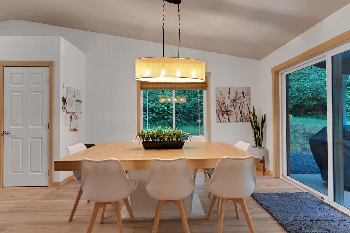 Dining area showcasing a light wood table with white chairs and a drum chandelier, featuring a white door to the left and large windows and a sliding glass door offering views of the outdoors.