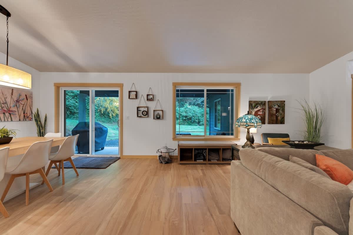 An open-concept living and dining area with light hardwood floors, featuring a dining table with chairs, a large sectional sofa, and a sliding glass door leading to the outdoor space.