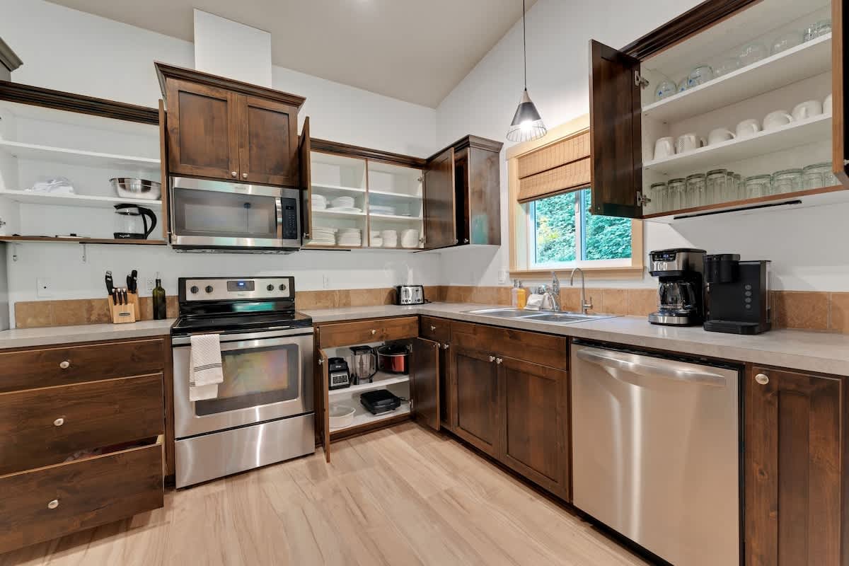 Modern kitchen with dark wood shaker-style cabinets, stainless steel appliances including a range, microwave, and dishwasher, and light beige countertops with a tile backsplash.