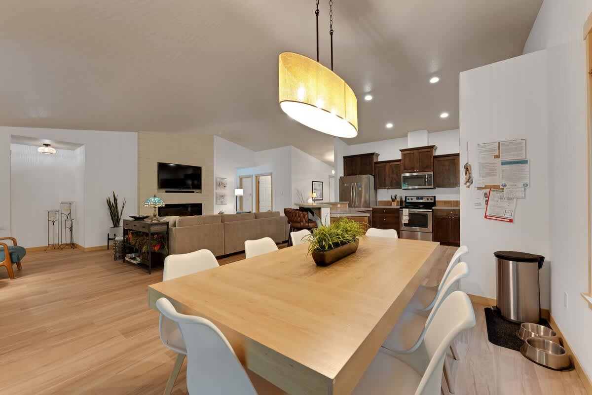 Open-concept dining area featuring a long light wood table with white chairs, a modern pendant light, seamlessly connecting to the kitchen and living room spaces.