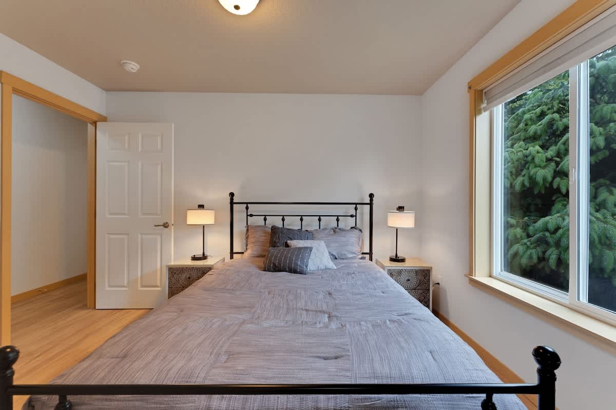 A cozy bedroom features a black metal frame bed with a gray patterned comforter and pillows, flanked by two nightstands with lamps. A large window on the right offers a view of lush green trees, while a white door leads to another room on the left.