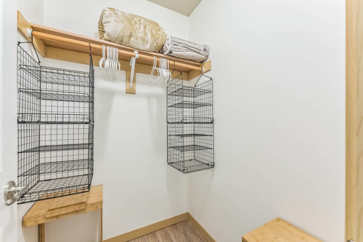 A well-organized closet features a light wood shelf with a hanging rod, holding blankets and white hangers. Black wire basket organizers are mounted on the walls, providing additional storage space.