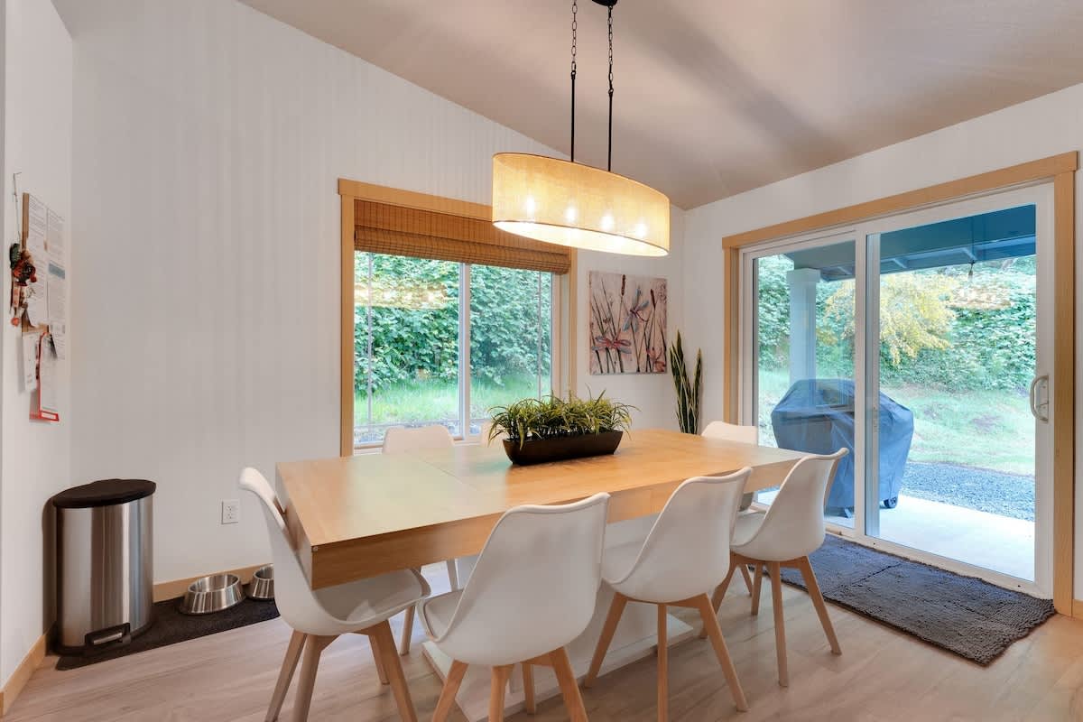 Bright dining area featuring a light wood rectangular table with six white modern chairs, illuminated by a drum-shaped chandelier, with large windows and a sliding glass door leading to an outdoor patio.