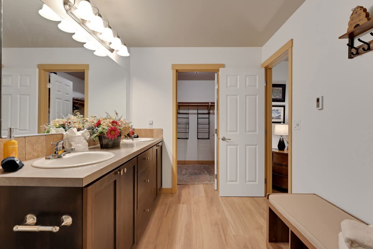 Spacious bathroom with a long double vanity featuring dark wood cabinets, light beige countertops, two sinks, a large mirror, and a view into a walk-in closet.
