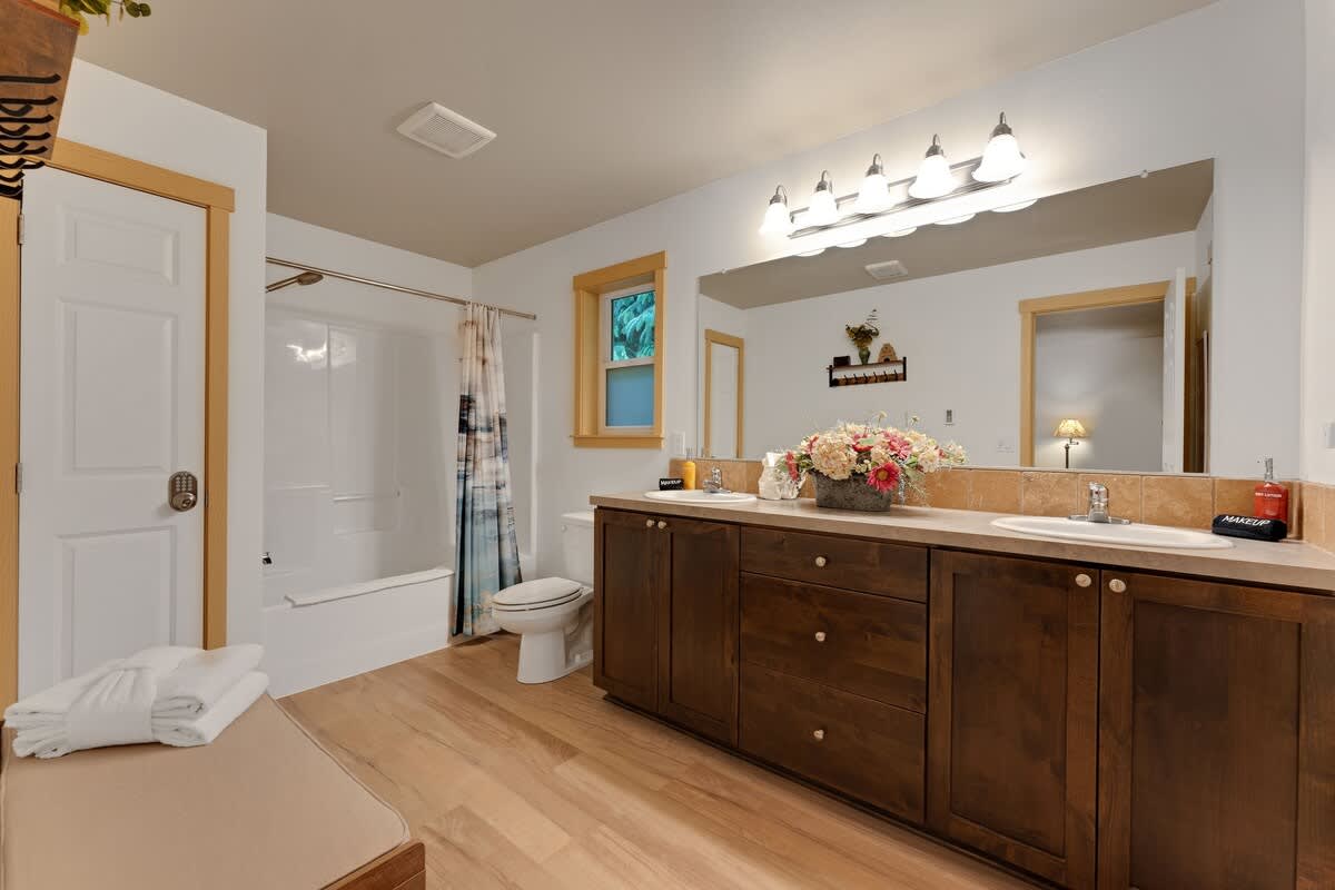 A spacious bathroom features a long dark wood double vanity with light countertops and two sinks, illuminated by a four-light fixture above a large mirror. A white bathtub with a shower and a toilet are visible on the left, with light wood flooring throughout.