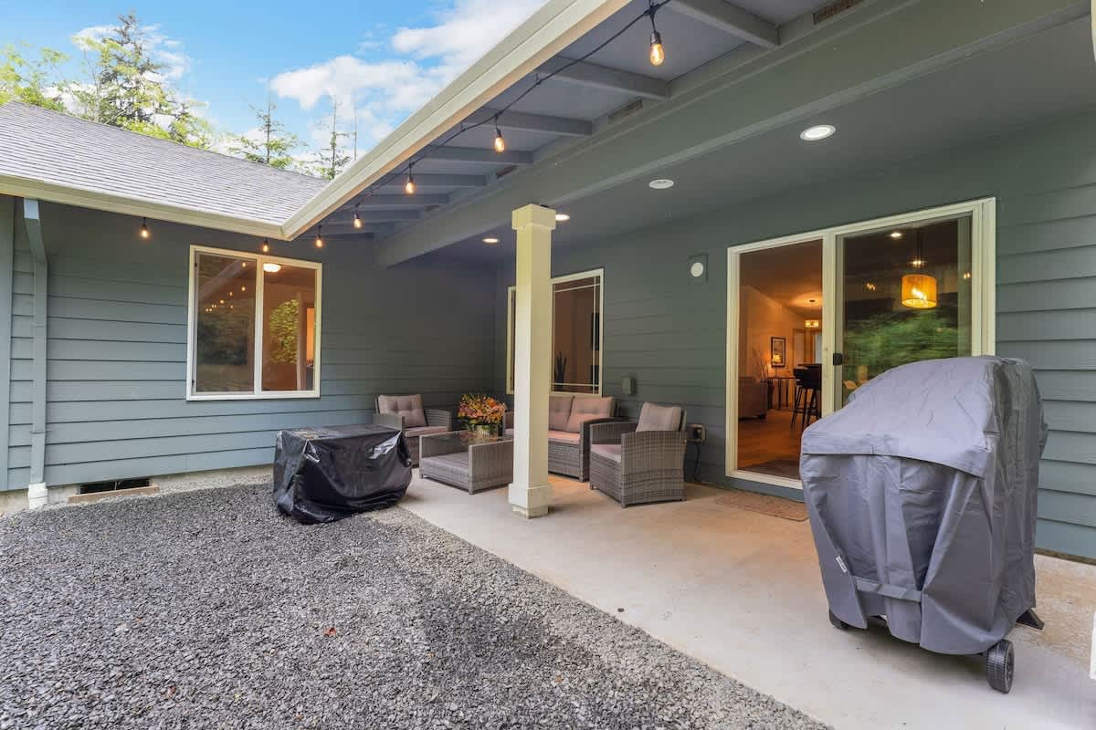 A concrete patio area with comfortable outdoor seating under a covered porch, featuring string lights and a covered BBQ grill, adjacent to a gravel path and the house exterior.