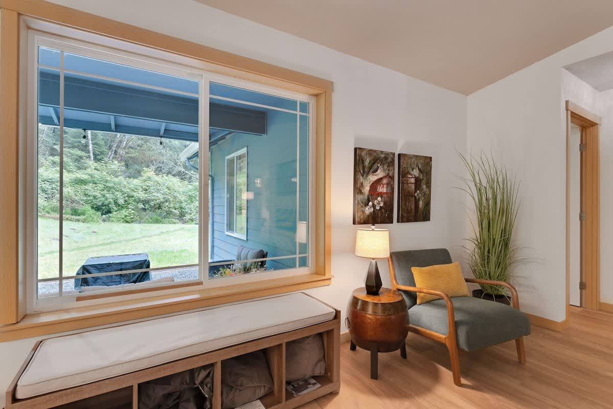 A cozy corner of the living room with a large sliding window framed in light wood, a window bench with storage, a teal armchair, and a barrel-shaped side table.
