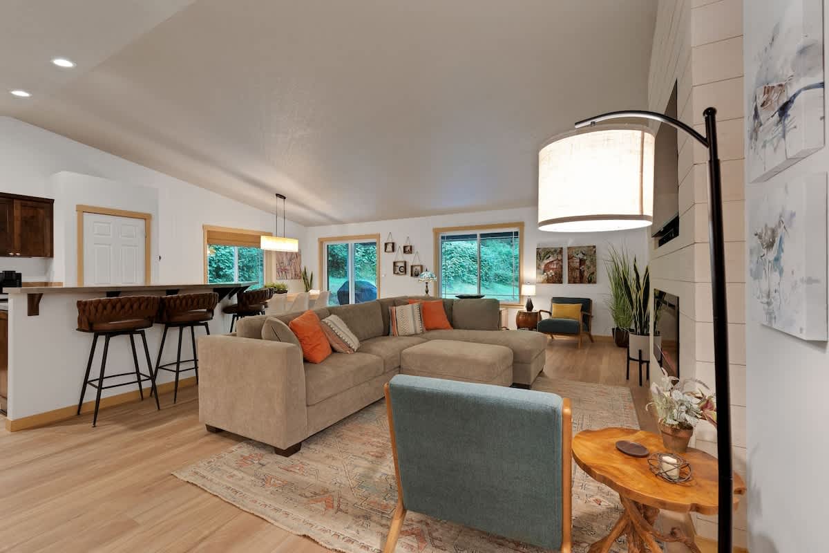 A bright and open living room with a large L-shaped sectional sofa and an area rug, facing a fireplace with a mounted TV, with a view of the kitchen bar and dining area.