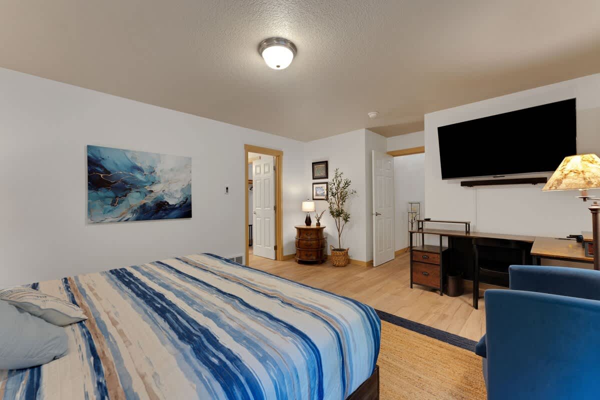 Bright bedroom featuring a queen bed with a blue and white striped duvet, a wall-mounted flat-screen TV, a dark wood desk with a chair, and a small nightstand with a plant.