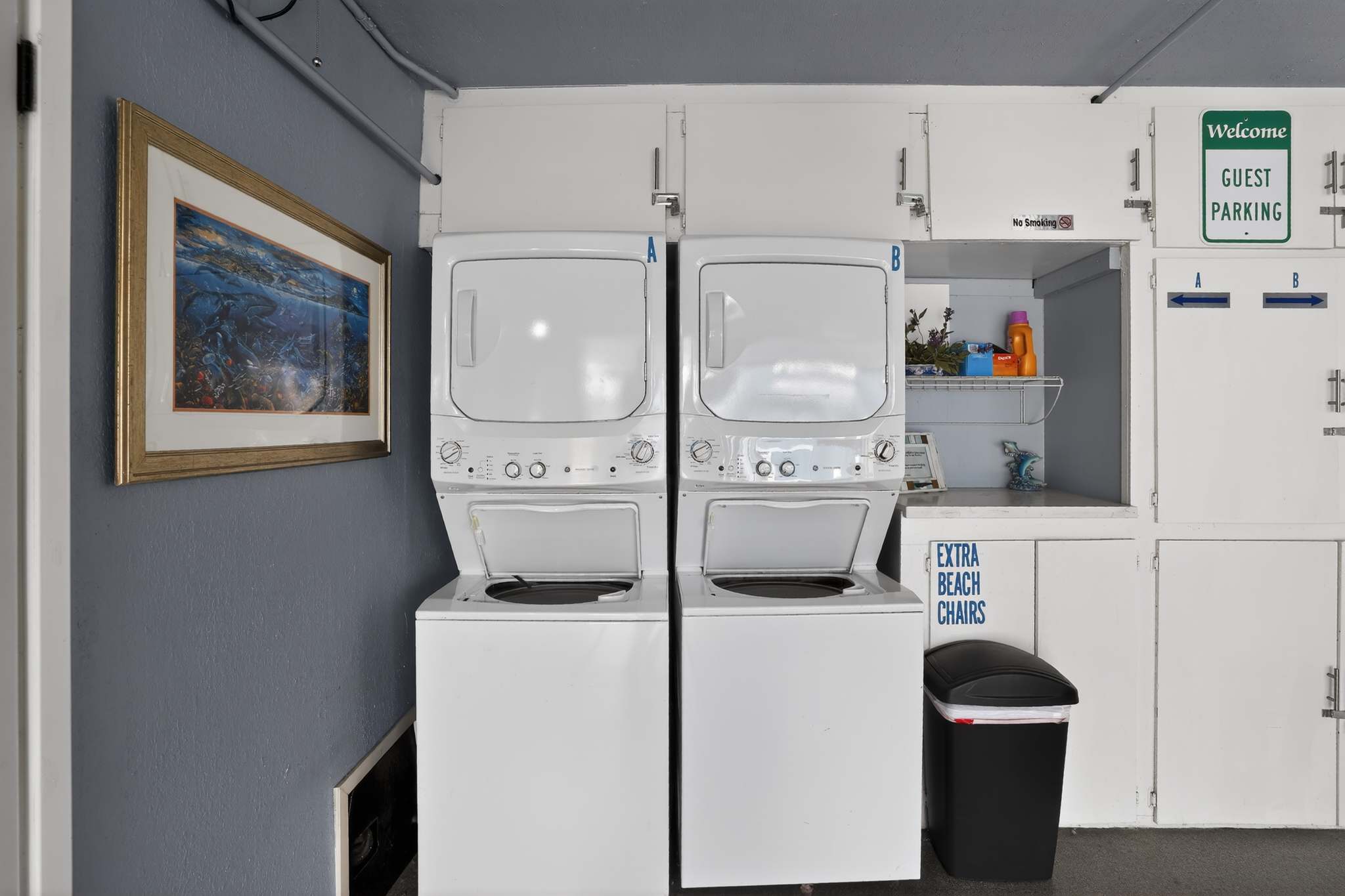 A functional laundry area features two stacked white washer and dryer units, a utility sink, and shelving with laundry supplies, alongside a "Guest Parking" sign.