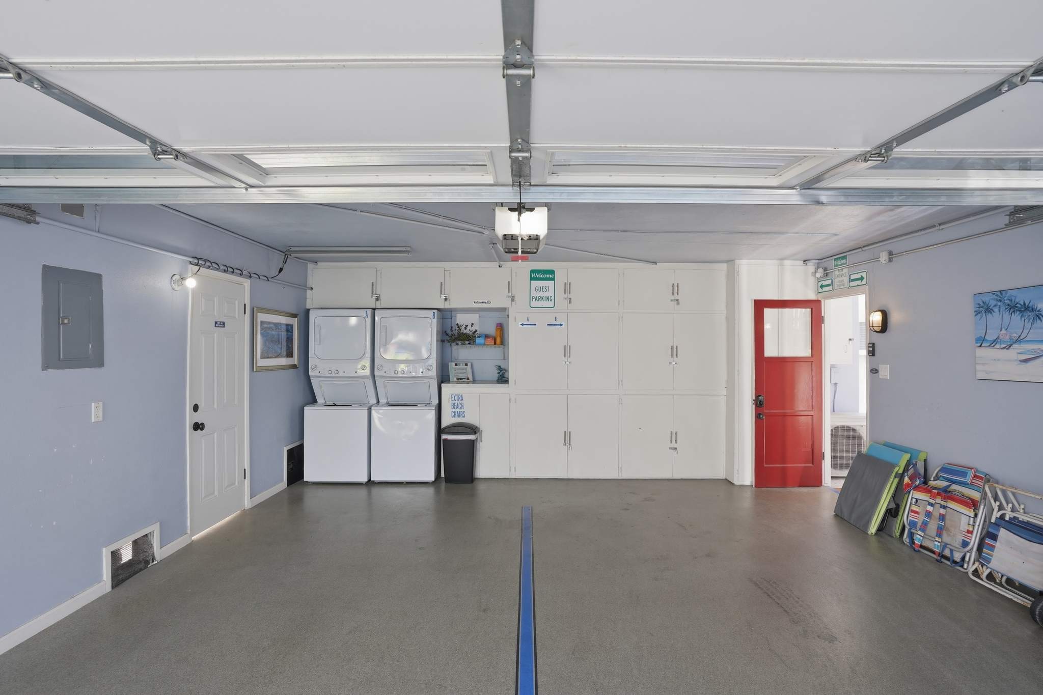 Spacious garage interior with light blue walls, a concrete floor, and a white washer and dryer set. Storage cabinets line one wall, and a red door leads to another area.