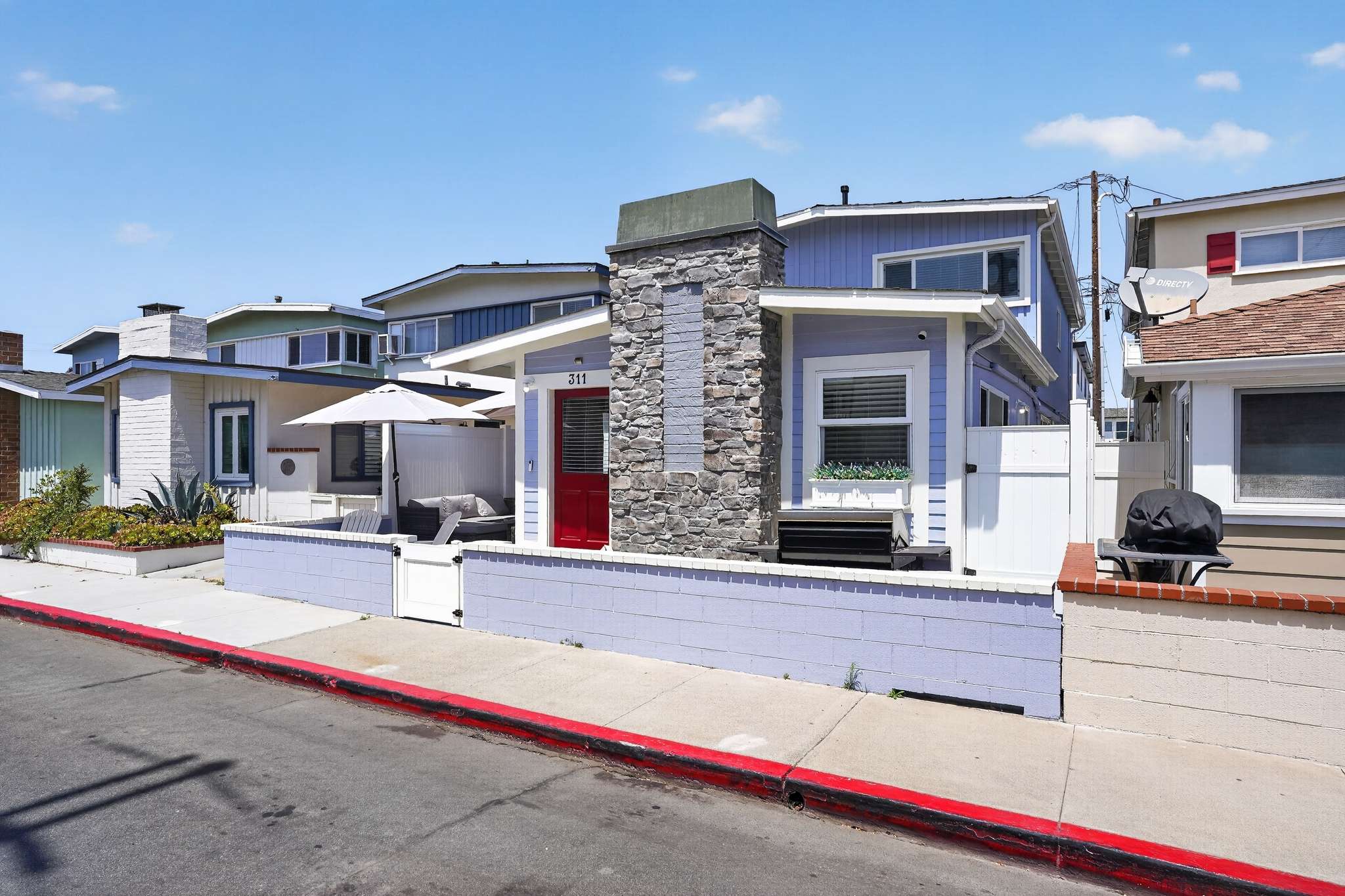 Front exterior of a light blue house with a red door and a stone chimney, featuring a small patio area enclosed by a low wall and white gate, facing a residential street.