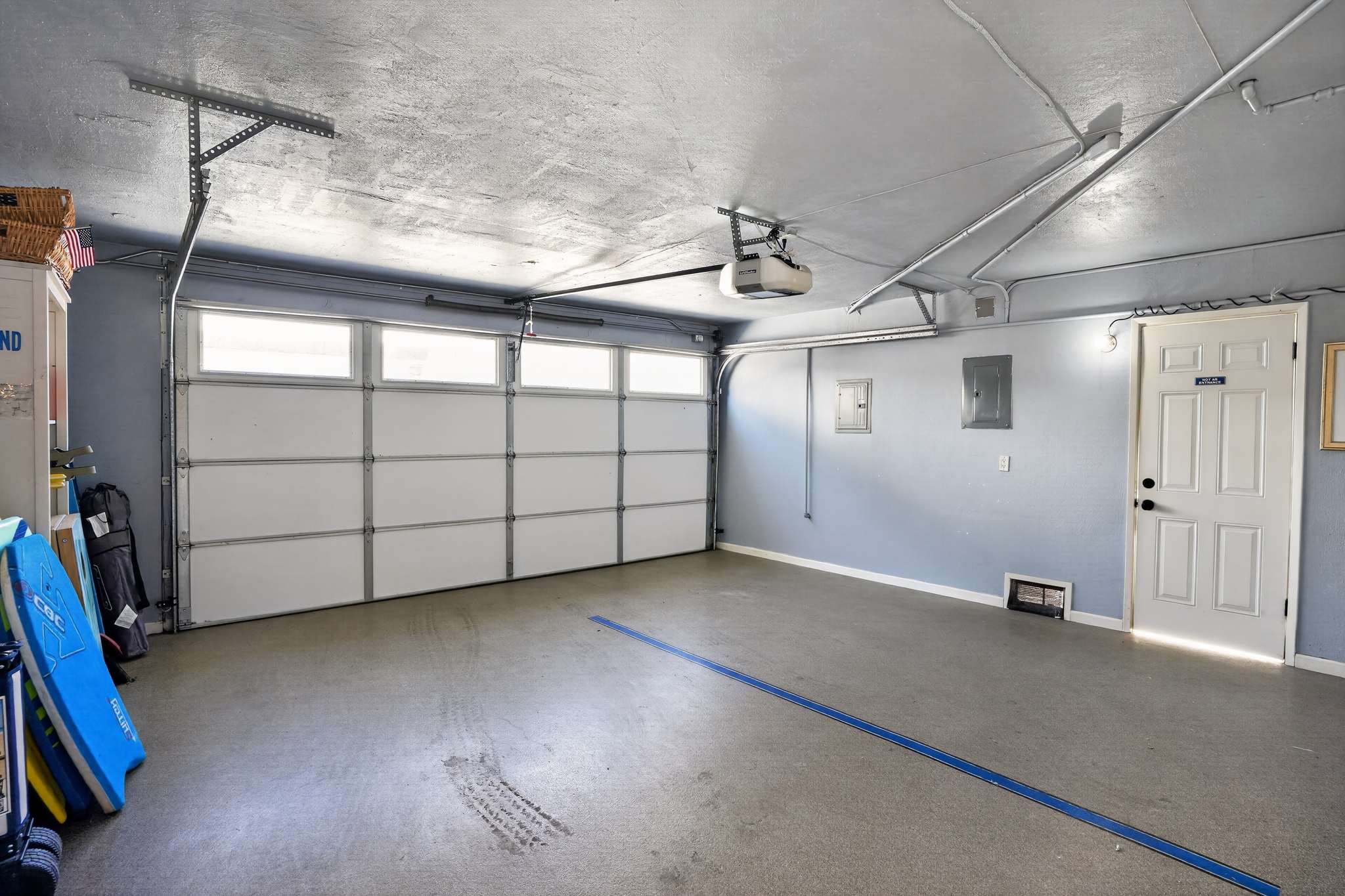 Interior view of a garage with a large white garage door, light blue walls, and a concrete floor. A side door is visible to the right, and an electrical panel is on the wall.
