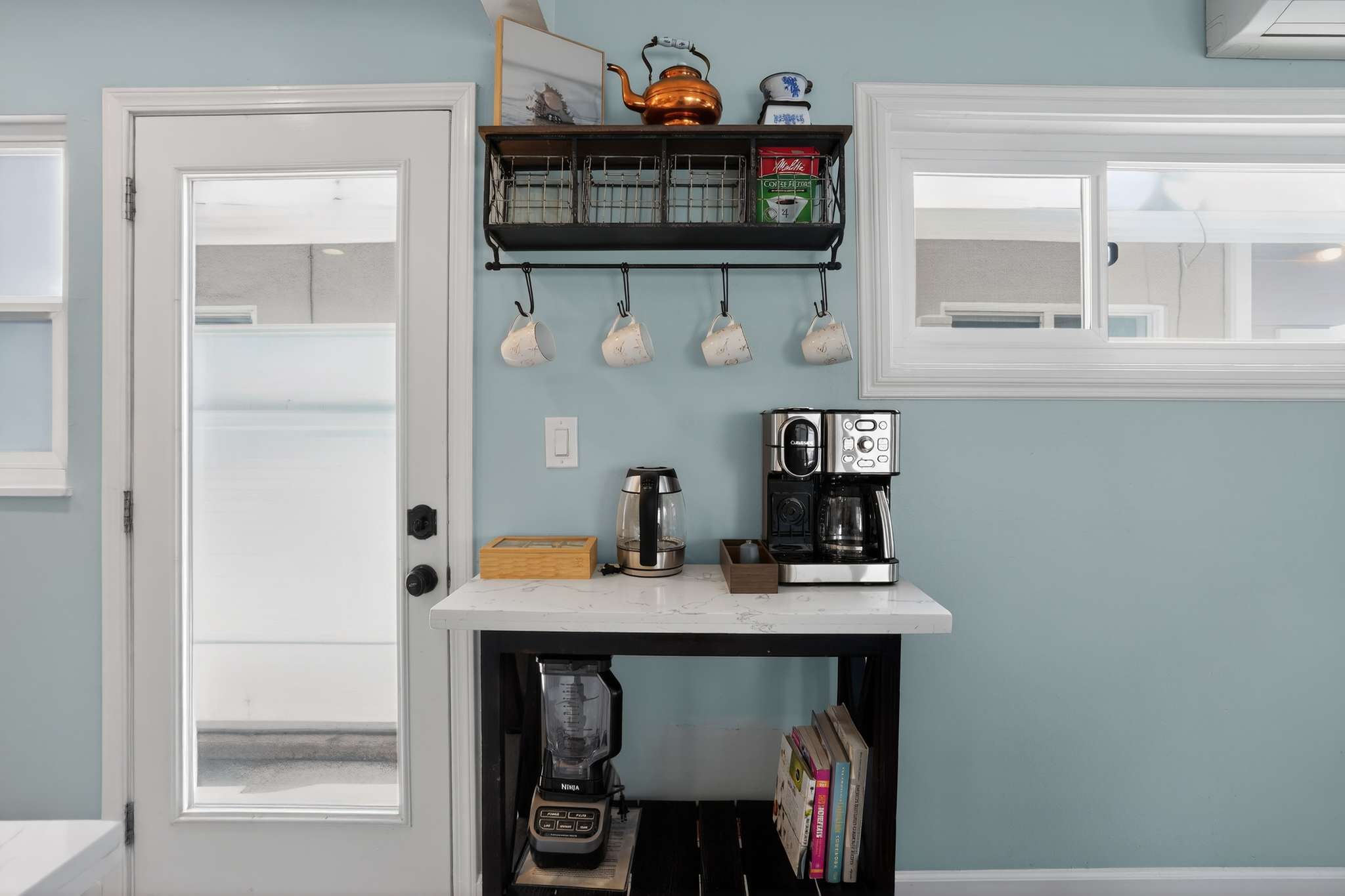 A dedicated coffee station is set up next to a white door, featuring a coffee maker, electric kettle, and a wall-mounted shelf with mugs and baskets, all against a light blue wall.