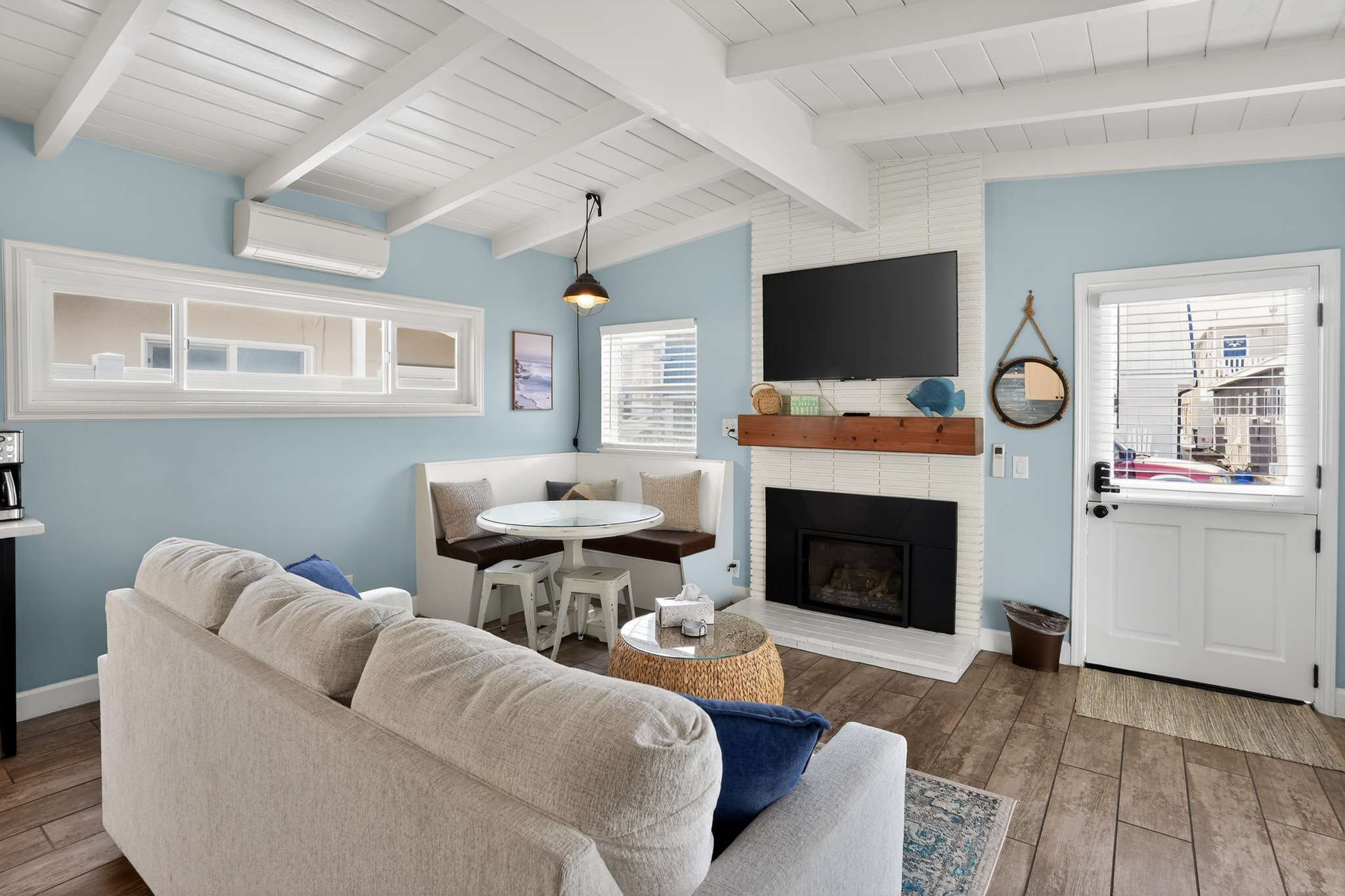 Living room featuring a white brick fireplace with a wooden mantel and a wall-mounted TV. A built-in dining bench with a round table is nestled in the corner, and a white dutch door is visible to the right.