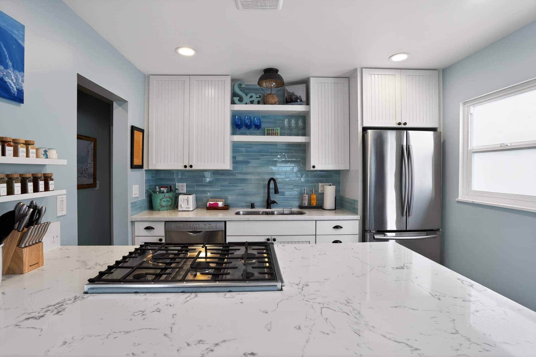 Modern kitchen with white shaker cabinets, light blue subway tile backsplash, and white quartz countertops. Stainless steel appliances, including a refrigerator and gas range, are visible.