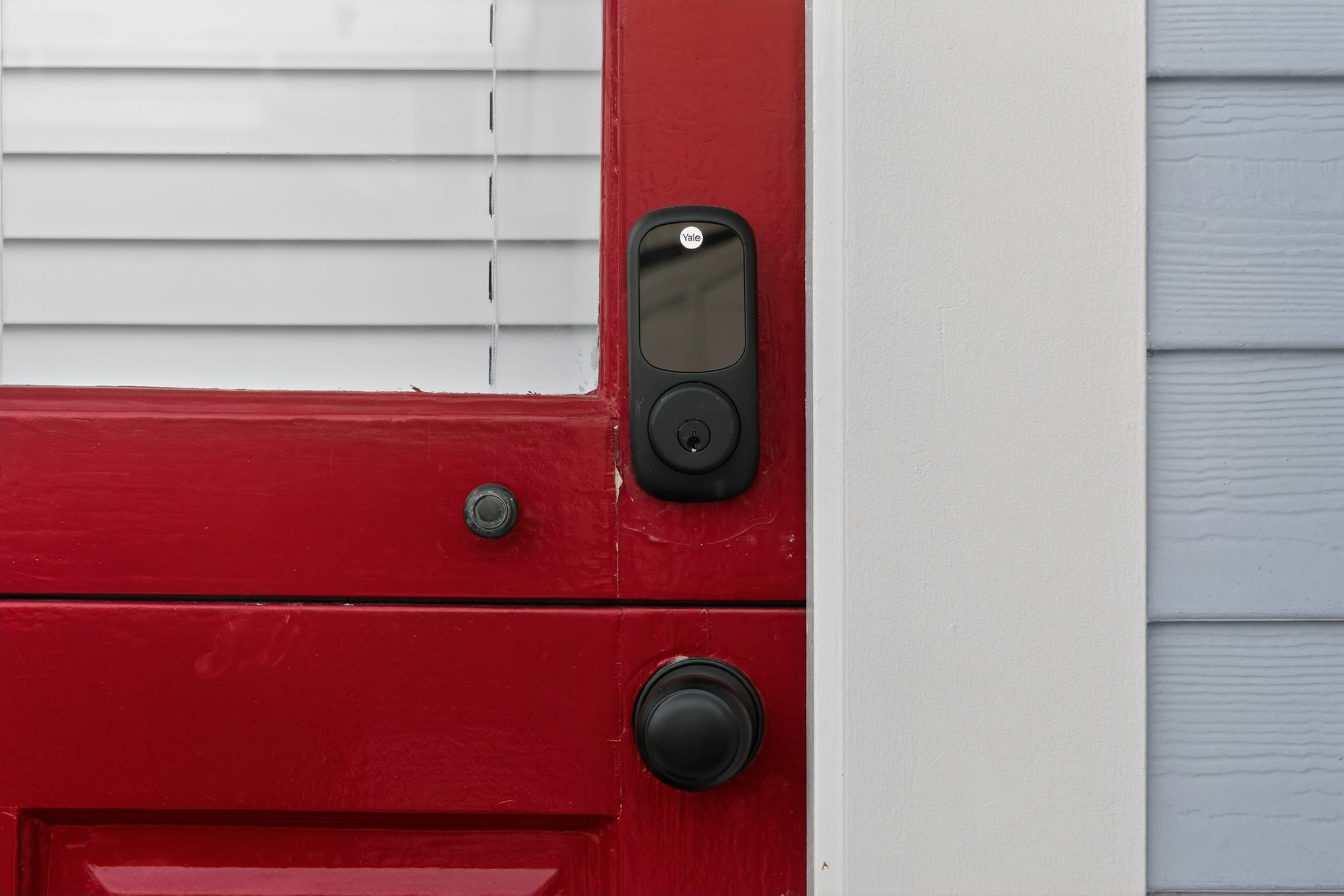 A close-up of a vibrant red entry door equipped with a modern black smart lock and a traditional black doorknob, set against light-colored siding.